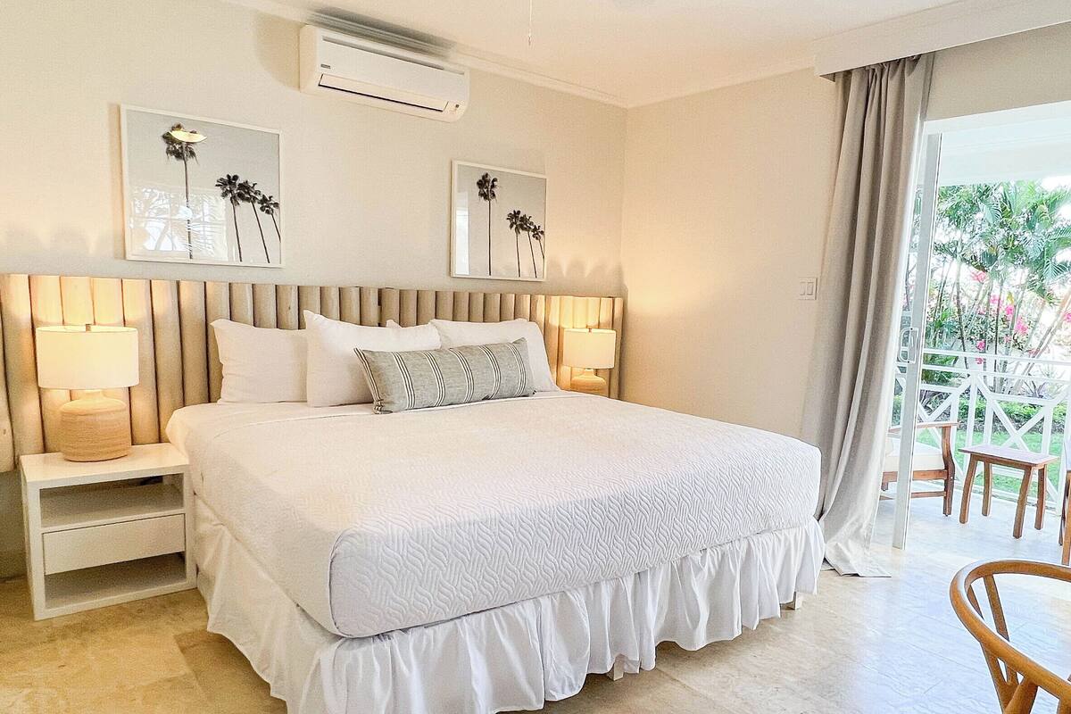 A hotel room with a large bed, two bedside tables with lamps, a ceiling air conditioner, and a view of a balcony with outdoor furniture.