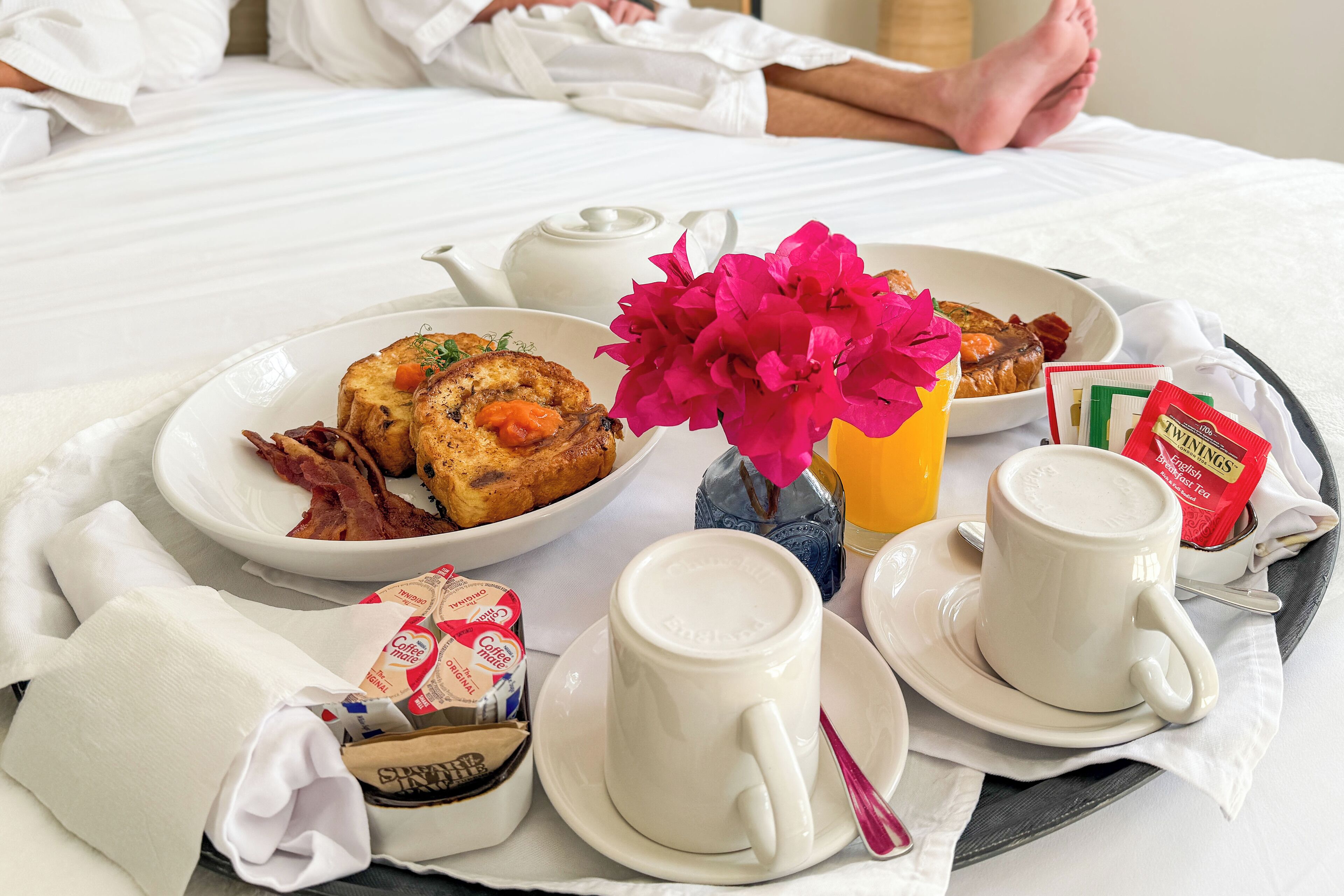 Bandeja de desayuno con tostadas, tocino, una flor, un hervidor de té y tazas de café sobre una cama.