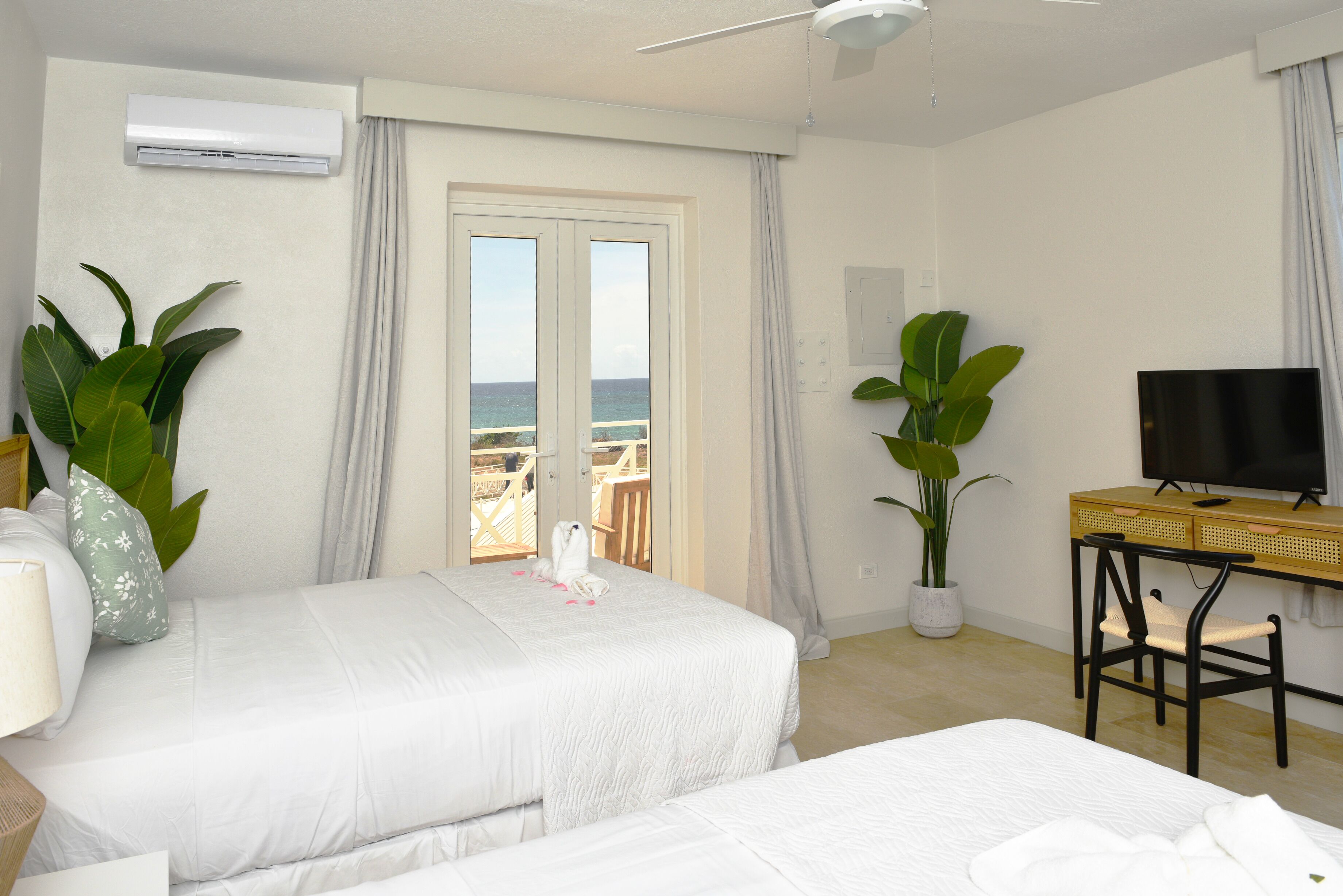 Habitación de hotel con dos camas, televisor, escritorio y grandes ventanales con vista al mar.