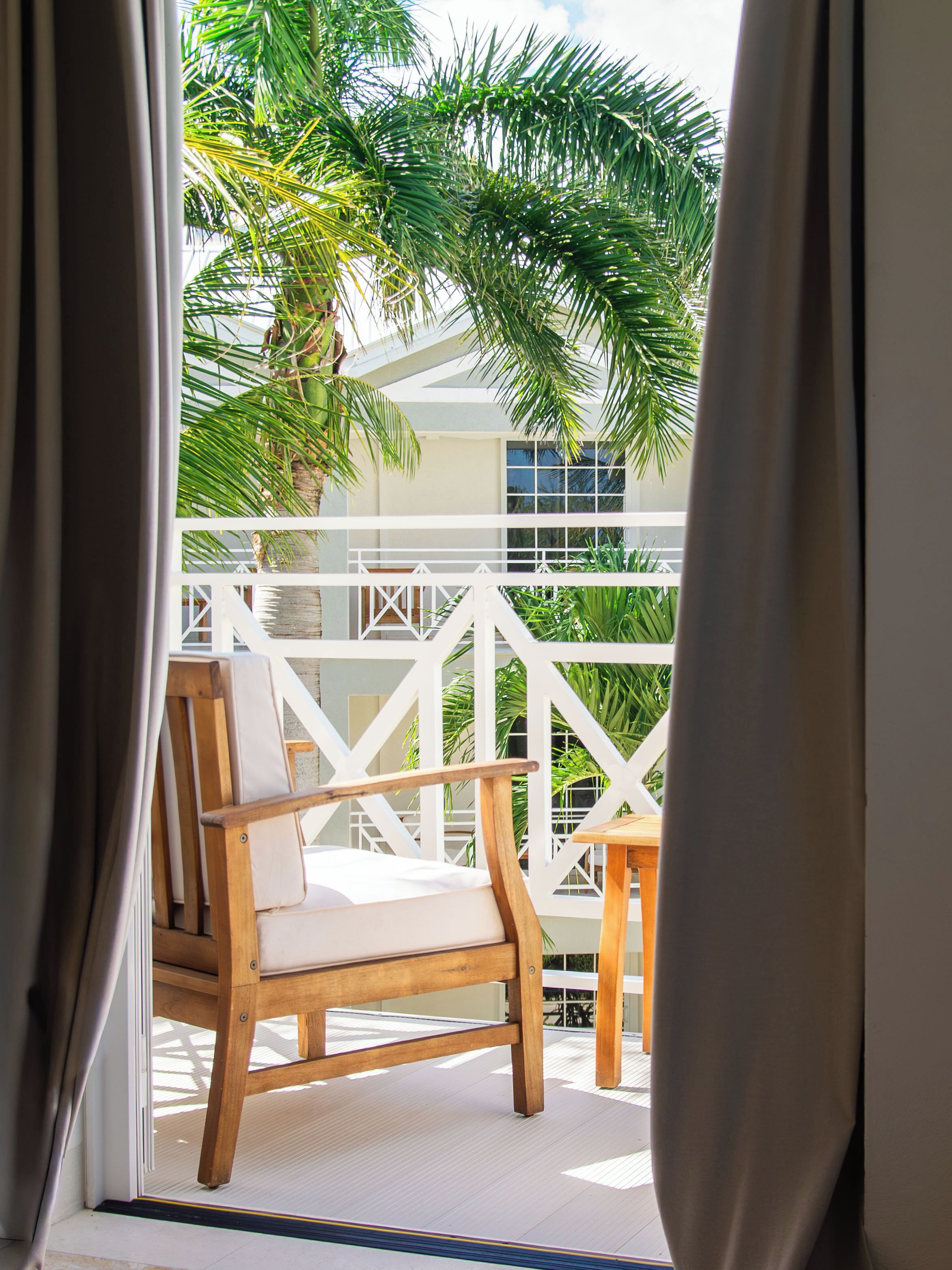 A wooden chair on a balcony with a white railing, overlooking a lush green landscape and a building with large windows.