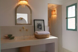 A bathroom with a white sink, a framed mirror, a small framed picture, a wicker basket with a towel, and a window with a green frame.