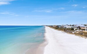 A coastal beach with white sand, clear turquoise water, and houses with balconies overlooking the ocean. - Sea Blue Escape | 30A 2BR | Pool + Walk to Beach (Santa Rosa Beach)