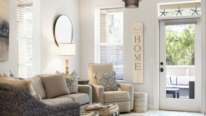 A living room with a sofa, armchair, and a door marked 'HOME'. - Sea Blue Escape | 30A 2BR | Pool + Walk to Beach (Santa Rosa Beach)
