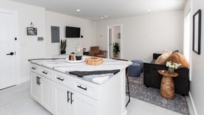 A modern kitchen with white cabinets, a marble countertop, and a built-in island. There is a flat-screen TV mounted on the wall, a potted plant, and a wooden side table with a vase. - Inn at Fulton: Unit 5 (Sandusky)