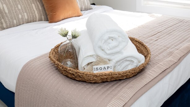 A neatly made bed with a rolled white towel, a woven basket with a glass container, and a label reading "SOAP CLEAN AS A WHISTLE". - Inn at Fulton: Unit 5 (Sandusky)