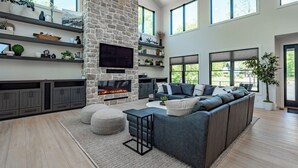 Ein geräumiges Wohnzimmer mit Kamin aus Stein, integrierten Regalen, einem großen grauen Ecksofa und großen Fenstern mit Blick ins Freie.