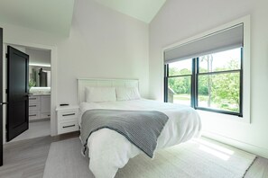 A modern bedroom with a large bed, white bedding, a gray throw, and a view of greenery through a large window.