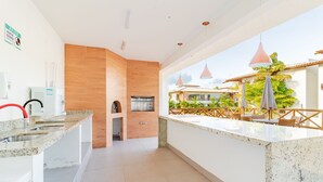 A modern kitchen with a marble countertop, a built-in oven, and a view of a pool area with outdoor seating. - IM05 Apto 3 Quartos - Riverside Imbassaí (Mata de São João)