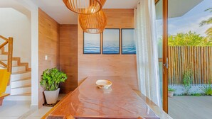 A modern interior with a wooden table, a potted plant, and a view of the sea through large windows. - IM05 Apto 3 Quartos - Riverside Imbassaí (Mata de São João)
