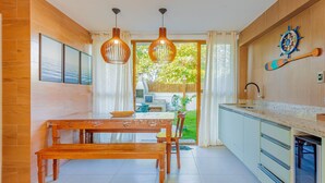 A modern kitchen with wooden cabinetry, a dining table with benches, pendant lights, and a view of the outdoors. - IM05 Apto 3 Quartos - Riverside Imbassaí (Mata de São João)