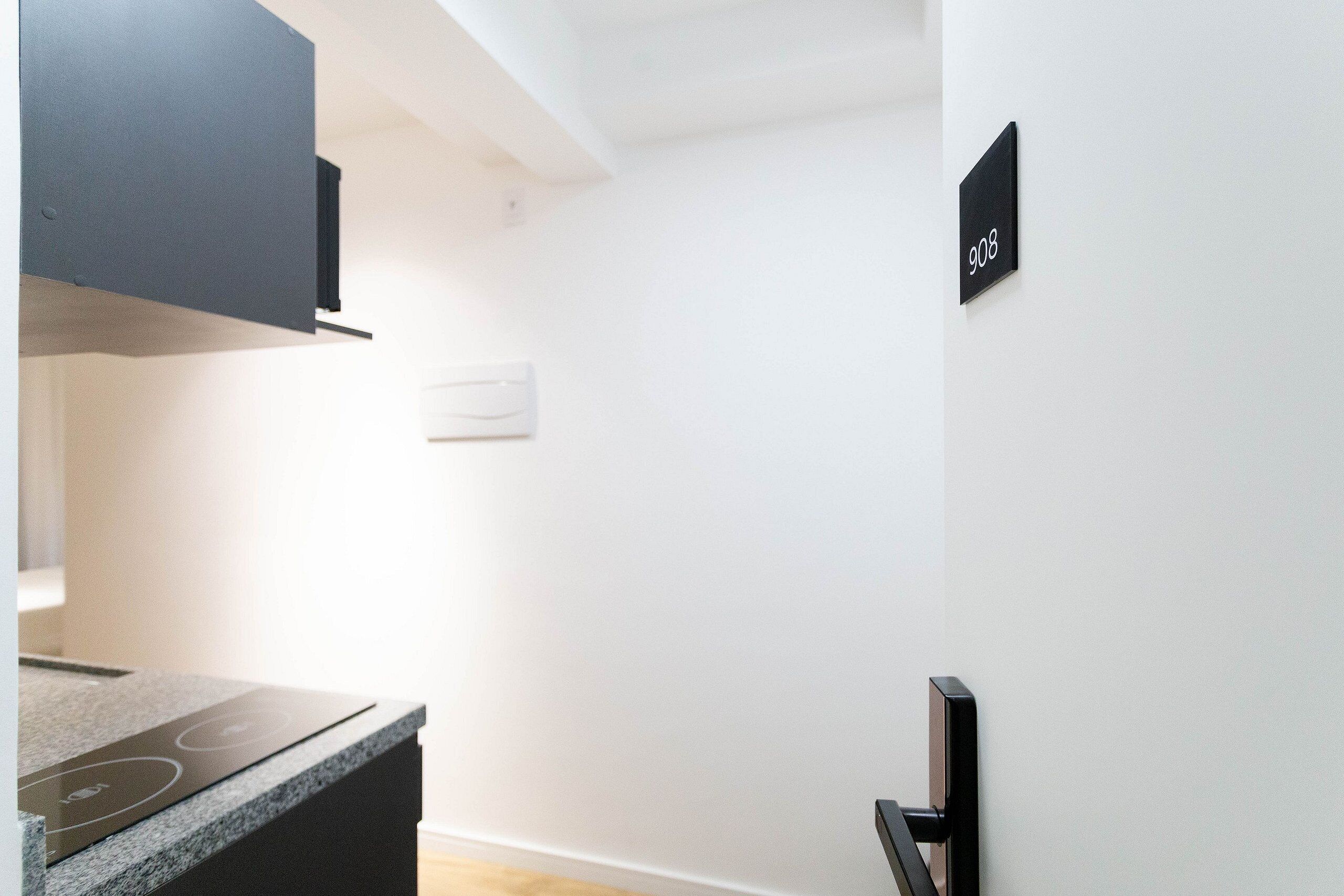 A modern kitchen with a built-in stove, a black cabinet, and a white wall with a black number plate indicating room 908.