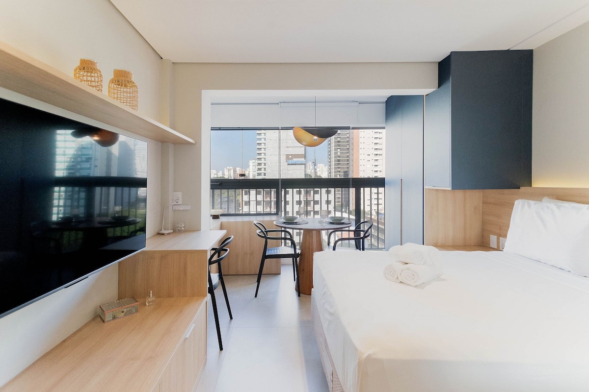 A modern hotel room with a large TV, a dining table with chairs, a bed with white linens, and a city view through large windows.