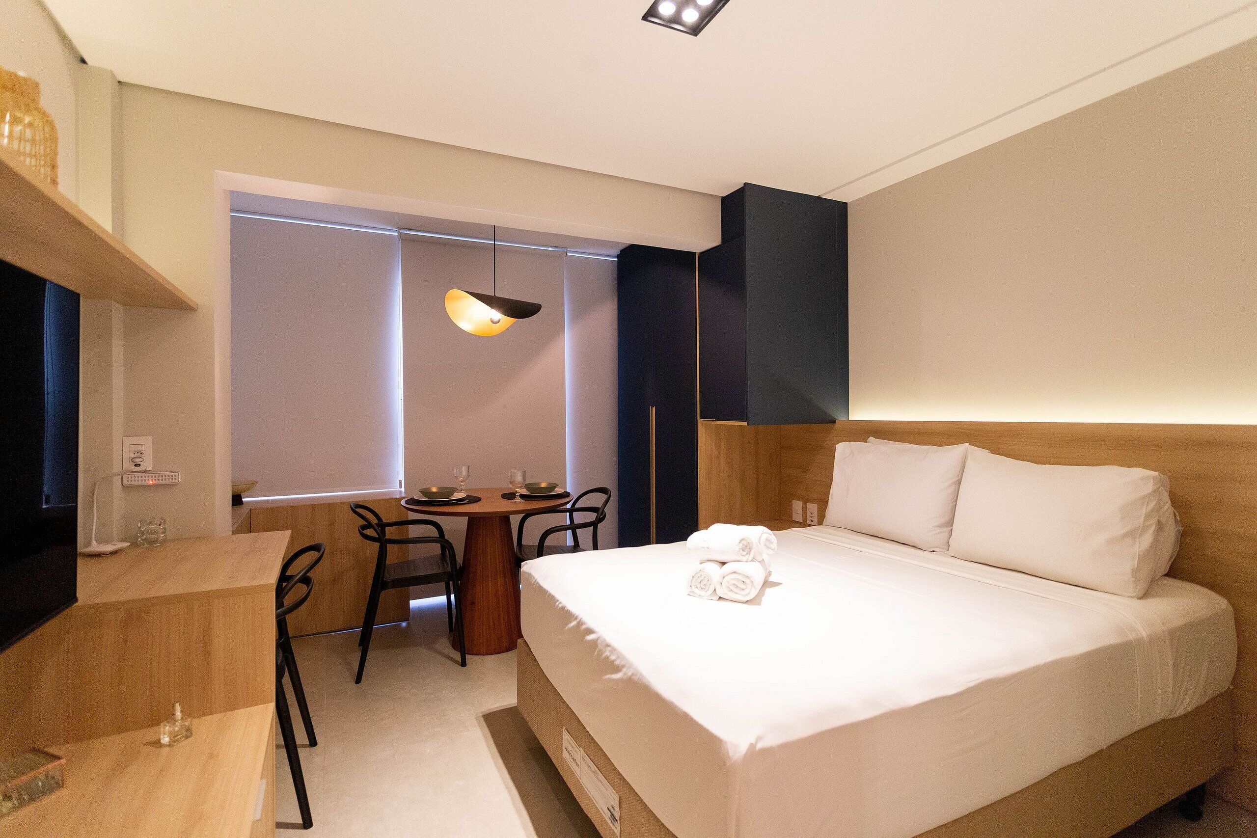 A hotel room with a bed, a desk, a chair, a table, and a window with blinds.