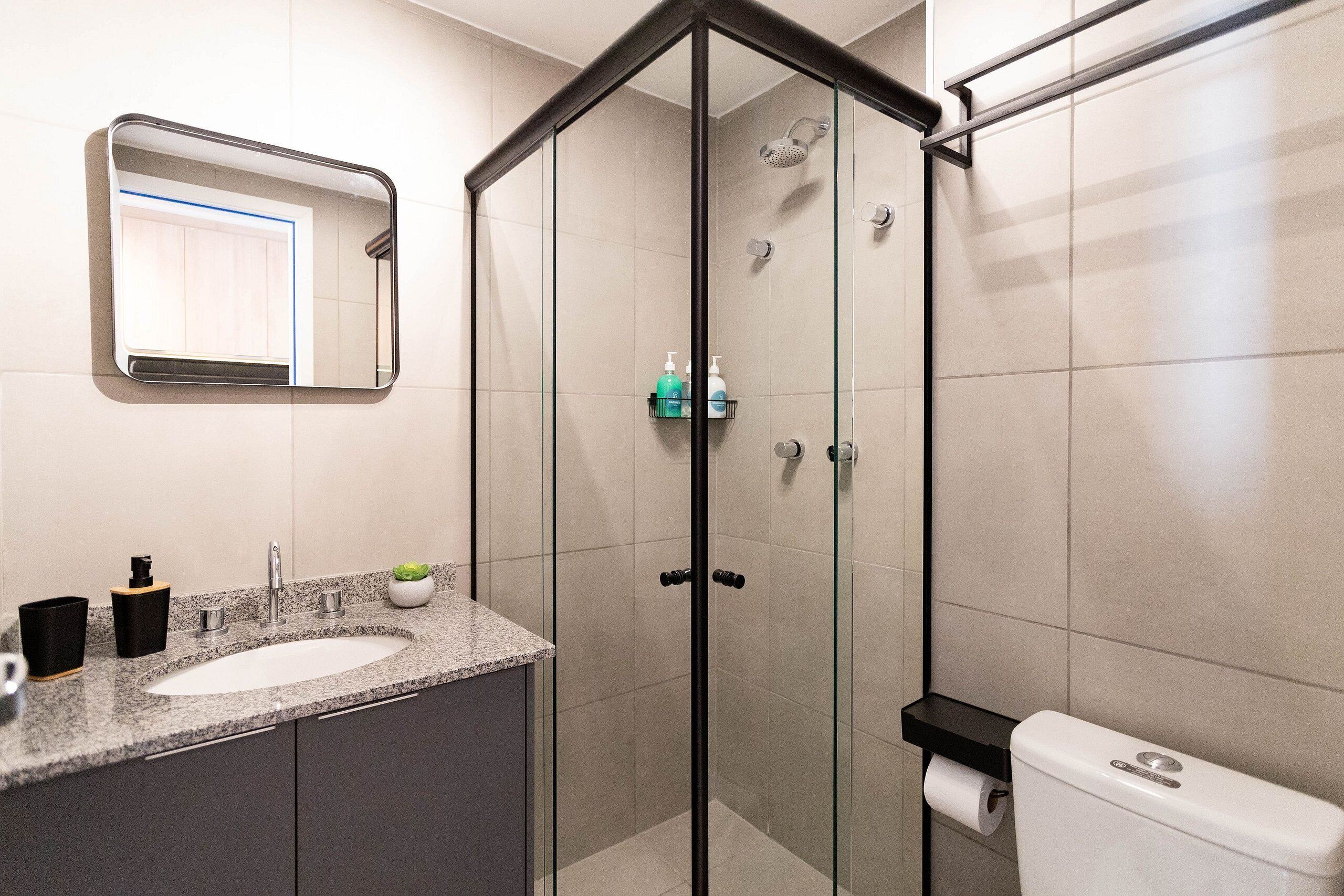 A modern bathroom with a glass-enclosed shower, a granite countertop sink, and a toilet with a paper towel holder.