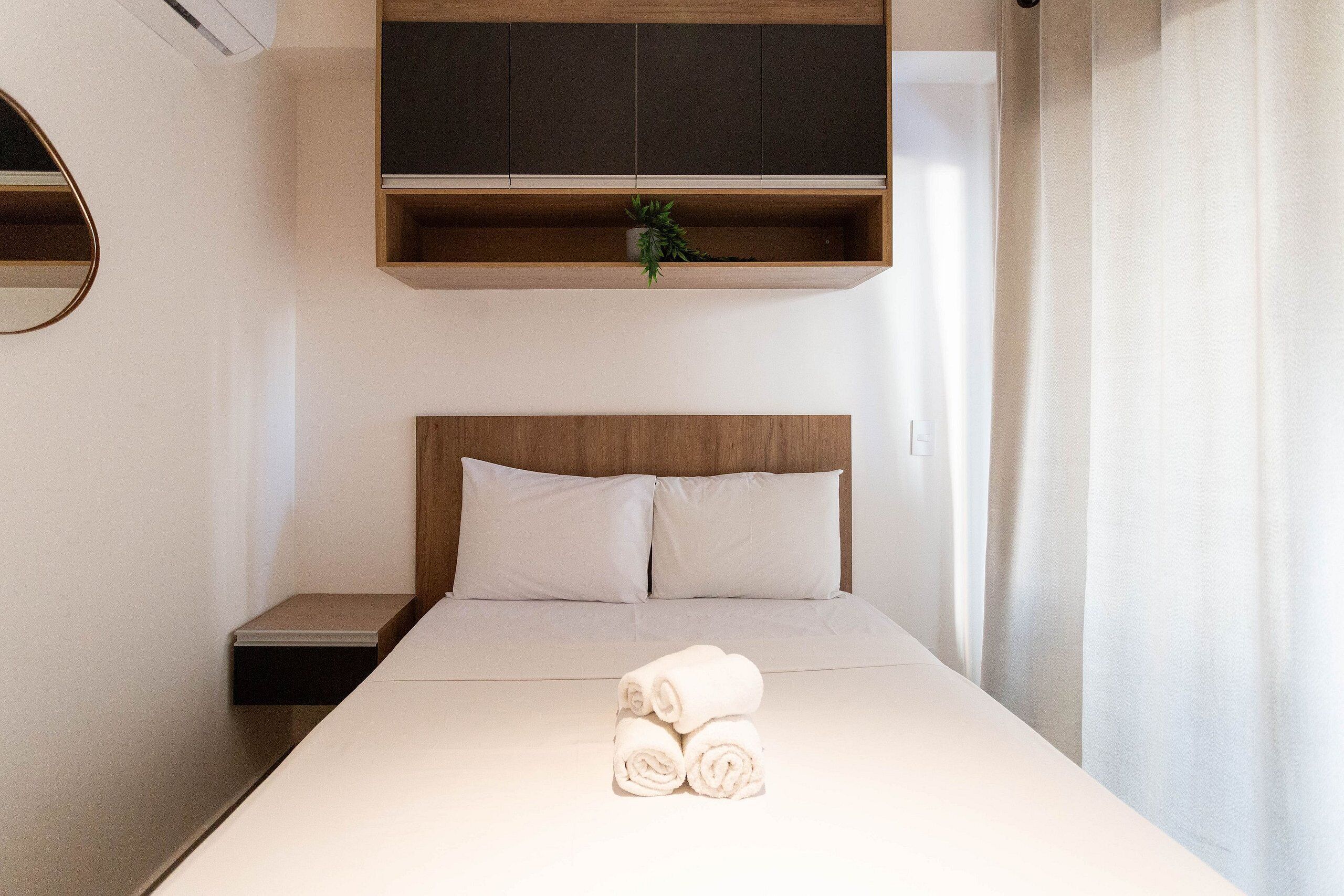 Un lit bien fait, avec des serviettes blanches pliées en rouleau, une tête de lit en bois et une petite étagère sur laquelle est posée une plante.