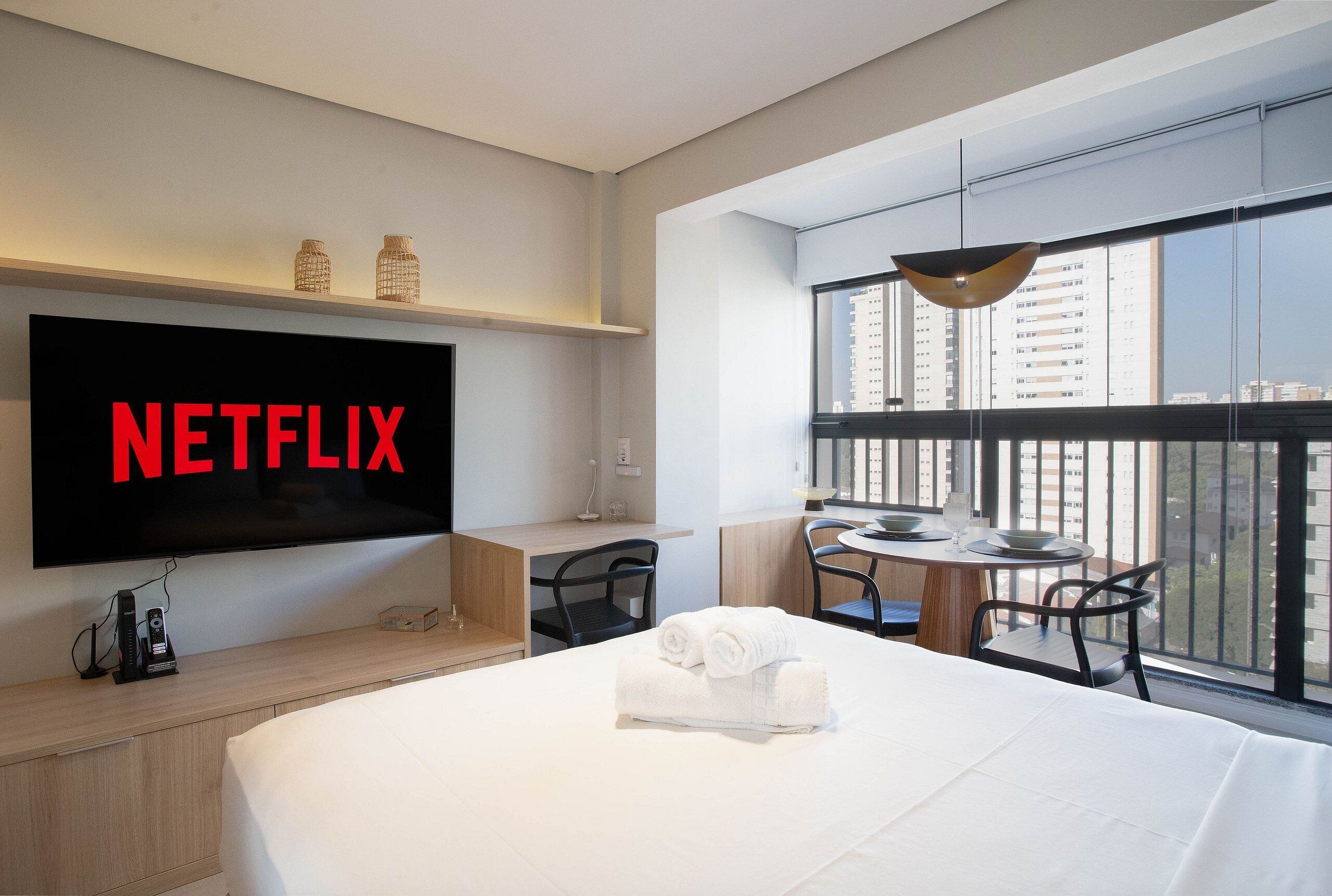 Habitación de hotel con una televisión de pantalla plana grande que muestra “NETFLIX”, una cama con toallas ordenadas, un escritorio con lámpara y un balcón con vista a la ciudad.