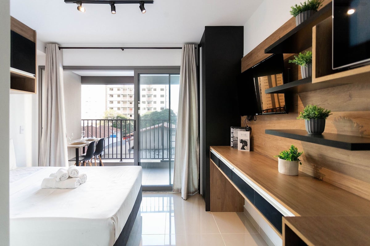 A modern hotel room with a bed, a desk, and a balcony with a view of the city.