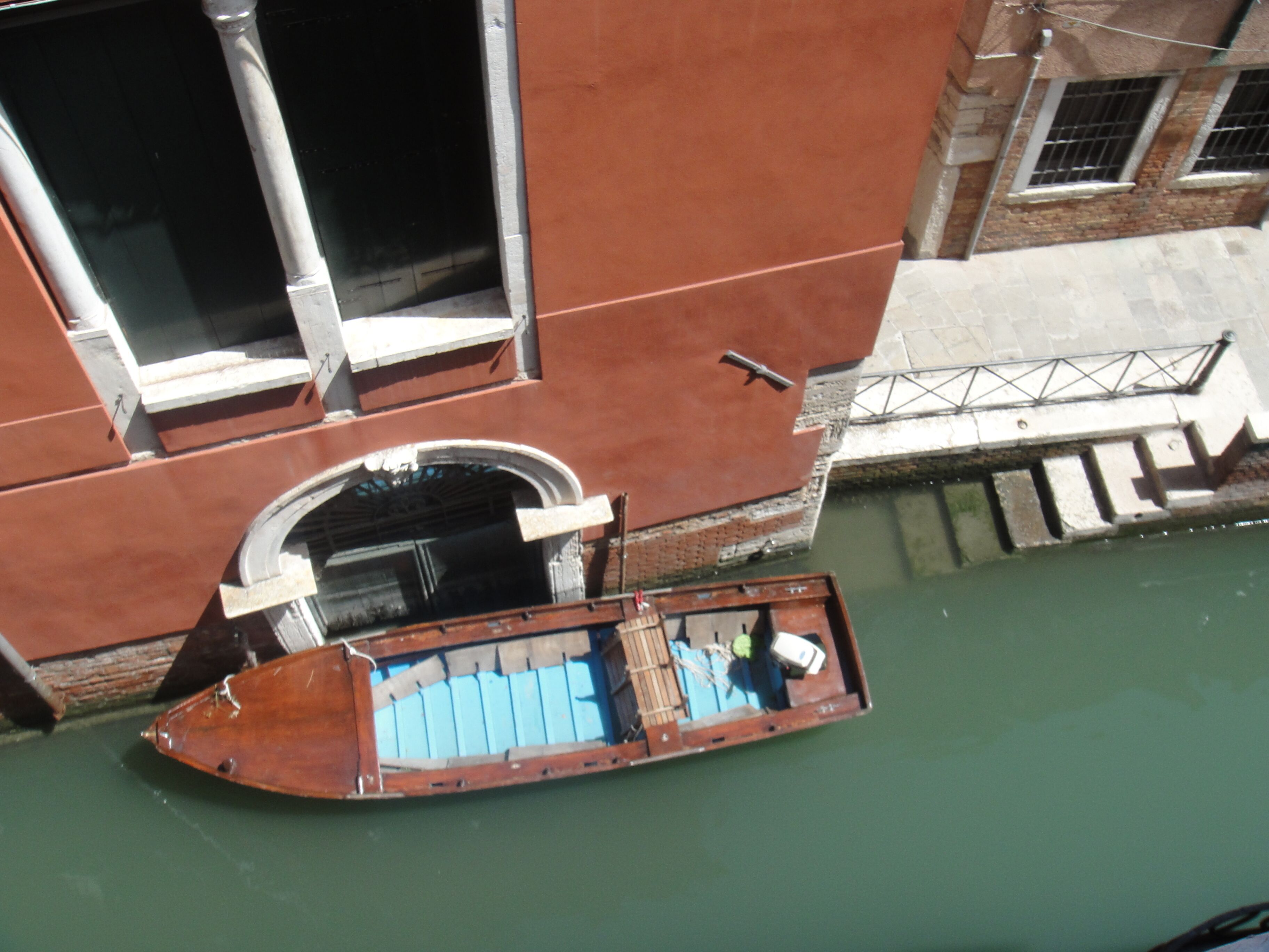 Un bateau à la coque en bois et aux sièges bleus est amarré dans un canal, devant un bâtiment rouge doté de grandes fenêtres et d’une rambarde en métal.