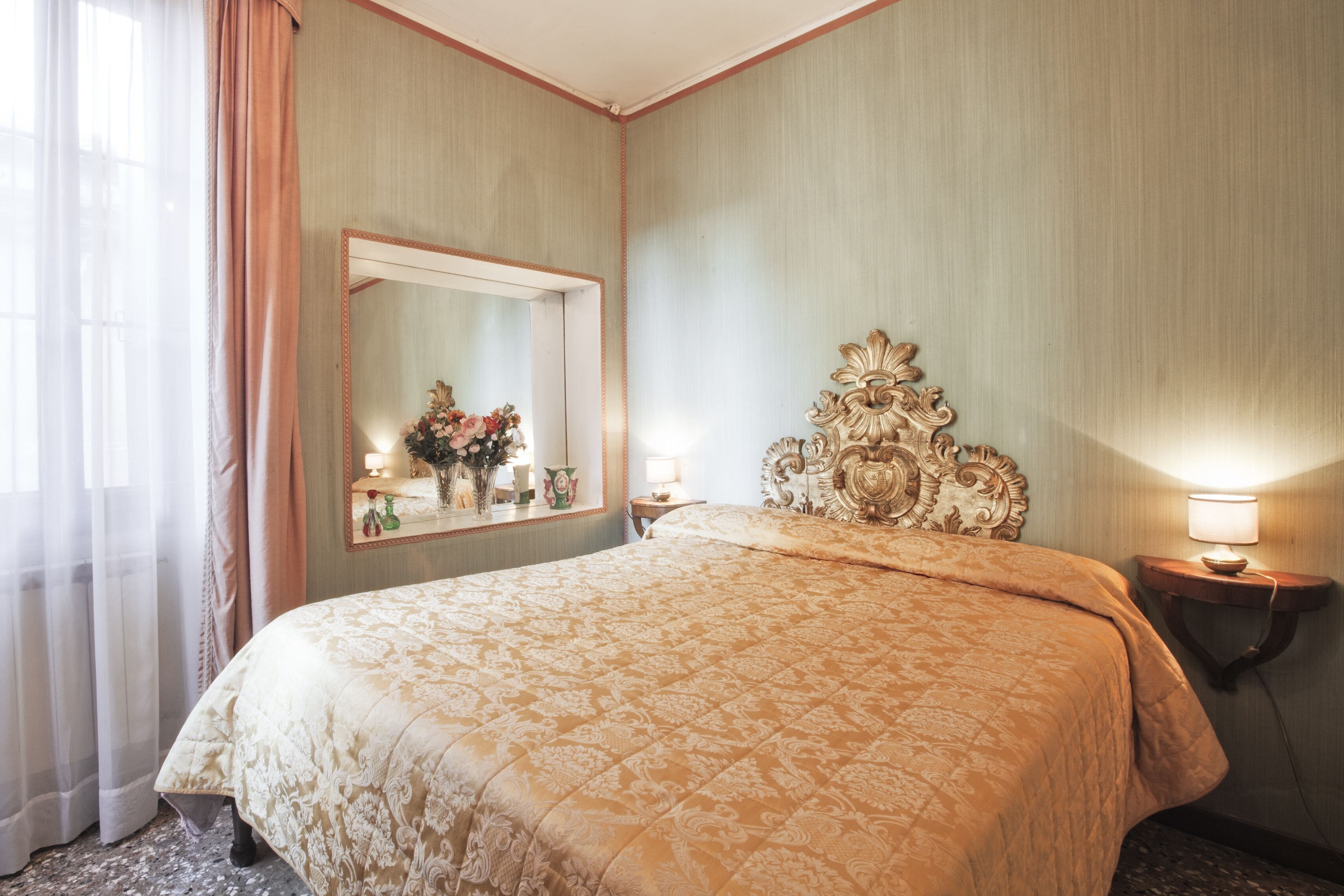 Habitación de hotel con una cama grande, un espejo, un cabecero decorativo y una mesita de noche con lámpara.