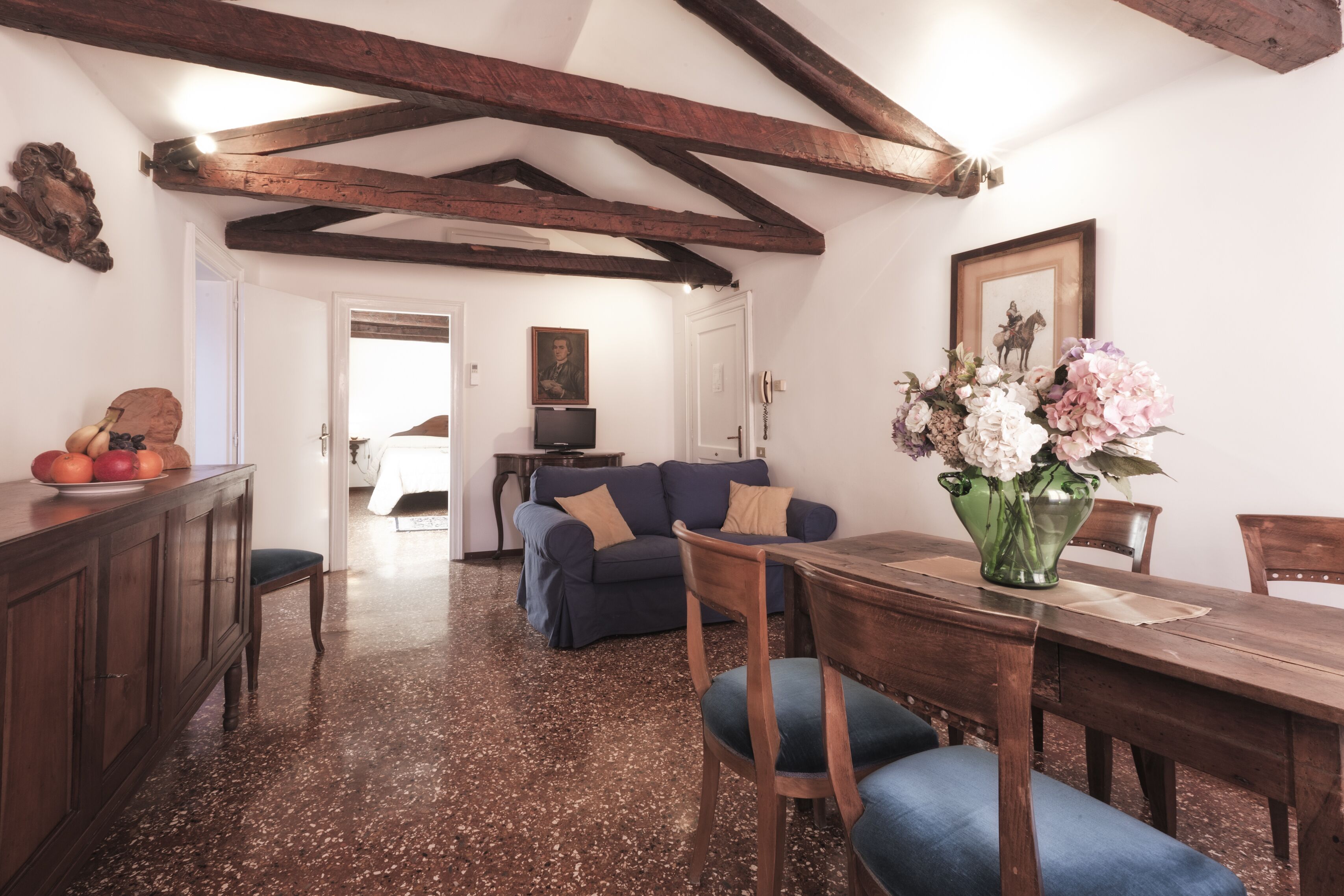 A dining area with a wooden table, chairs, and a vase of flowers. There is a sofa and a television in the background.
