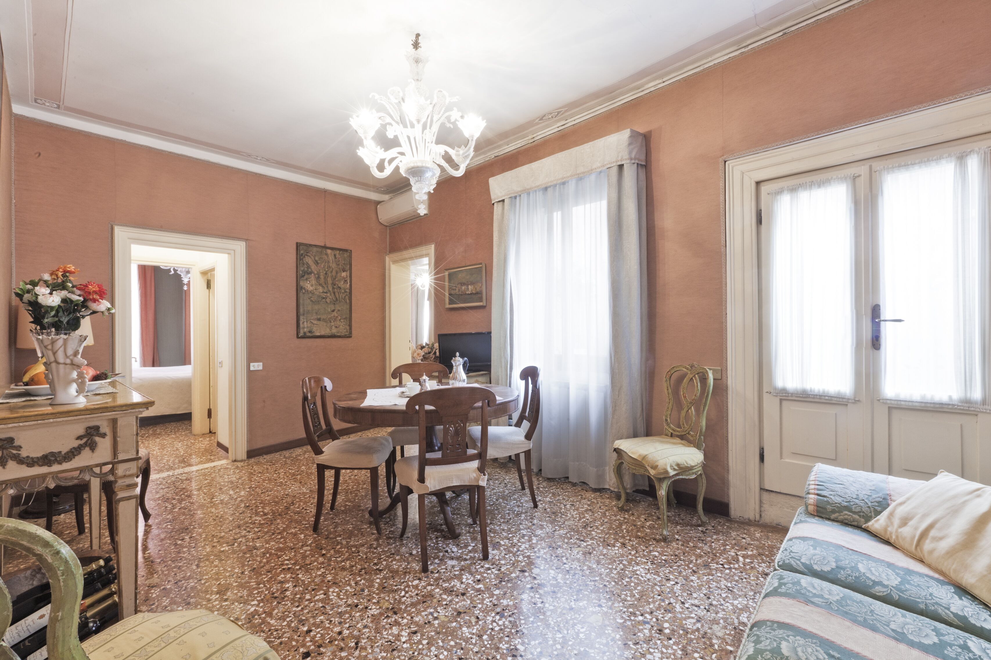 A dining area with a table, chairs, and a chandelier. There is a sofa and a window with curtains.