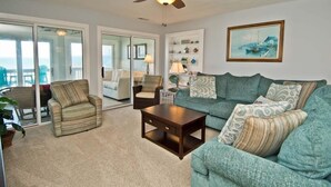 A spacious living room with a large sofa, a coffee table, and two armchairs. There is a ceiling fan, a painting on the wall, and a view through the glass doors. - Sun Bay 6 - Oceanfront Condo in Atlantic Beach, NC! (Pine Knoll Shores)