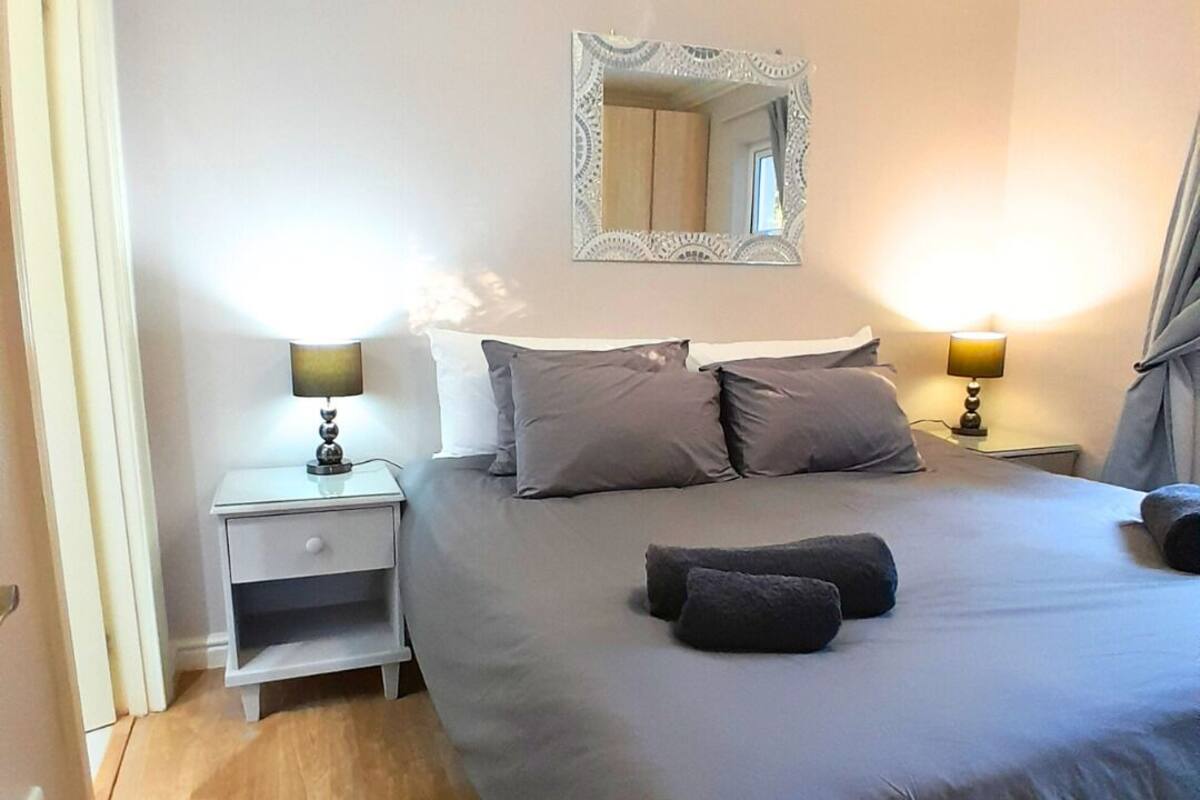 A neatly arranged bedroom with a large bed, two bedside tables with lamps, a mirror above the bed, and a wooden floor.