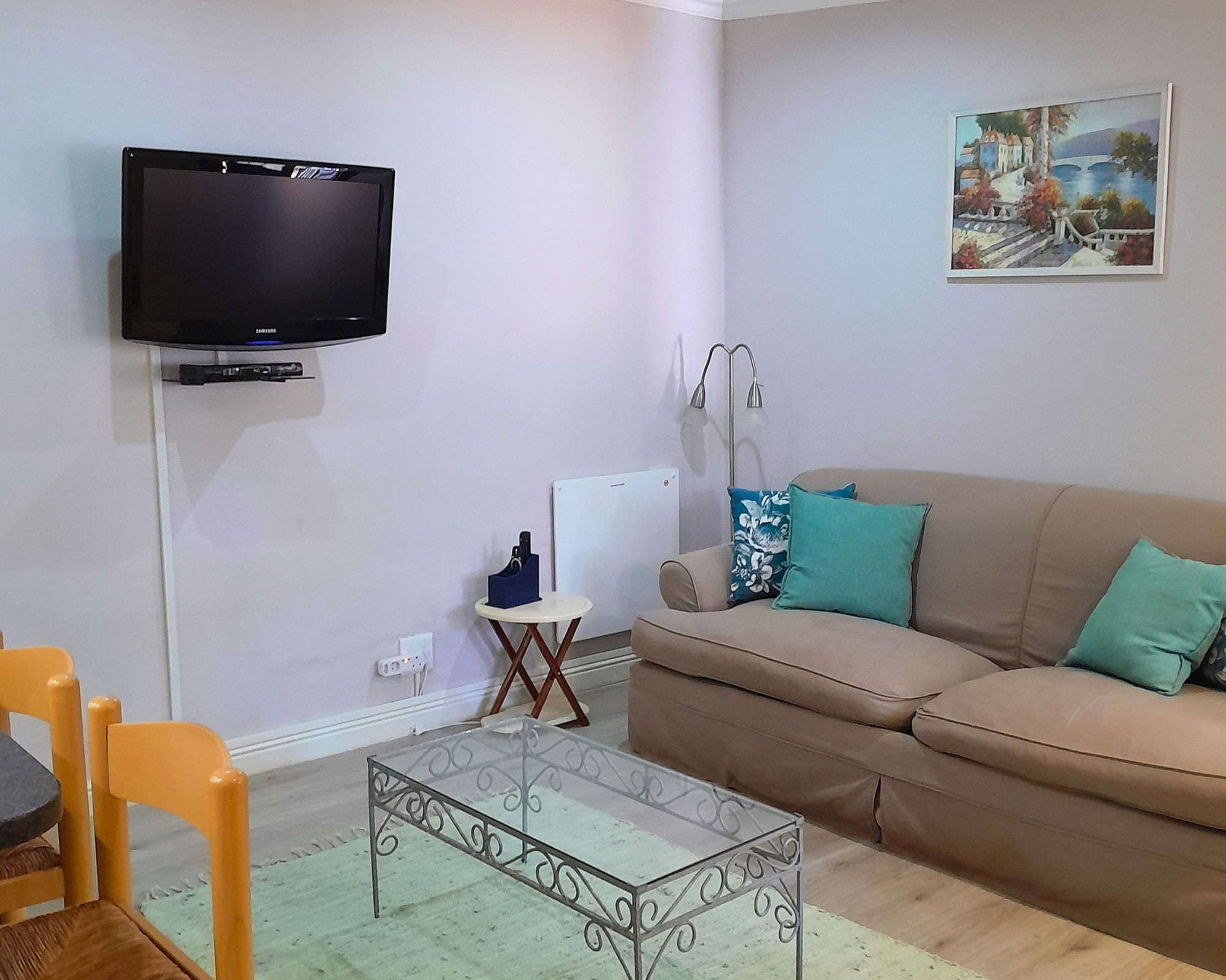 Ein Wohnzimmer mit an der Wand montiertem Fernseher, einer Couch mit Kissen, einem kleinen Tisch und einem Sessel.