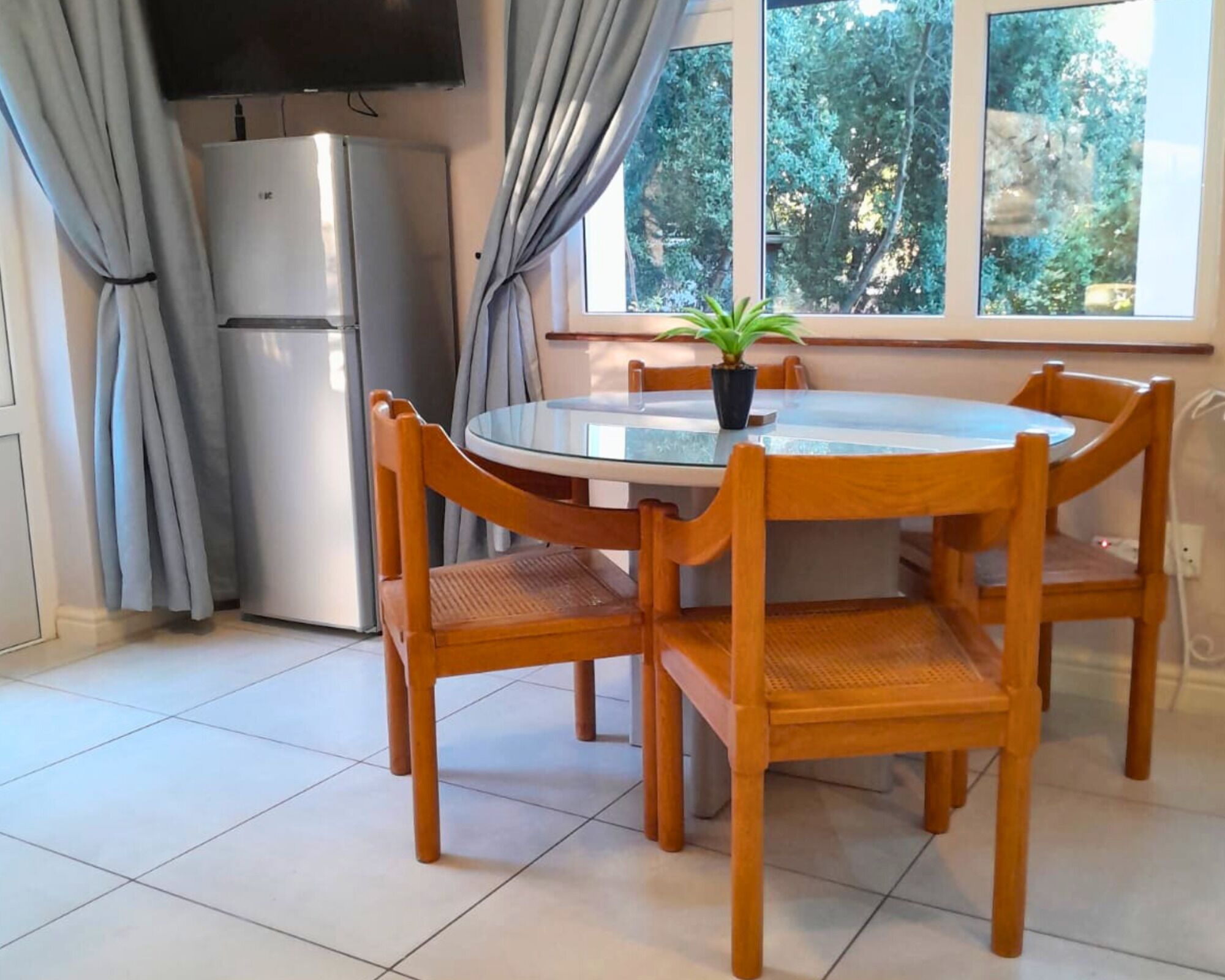 A dining area with a round glass table, four wooden chairs, a potted plant, a refrigerator, and a window with curtains.