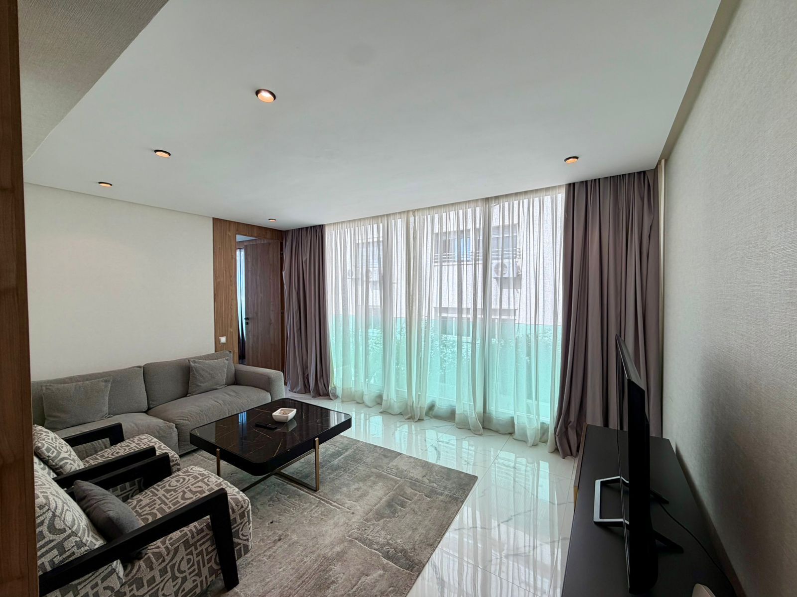 A modern living room with a sofa, a coffee table, a flat-screen TV, and large windows with curtains.