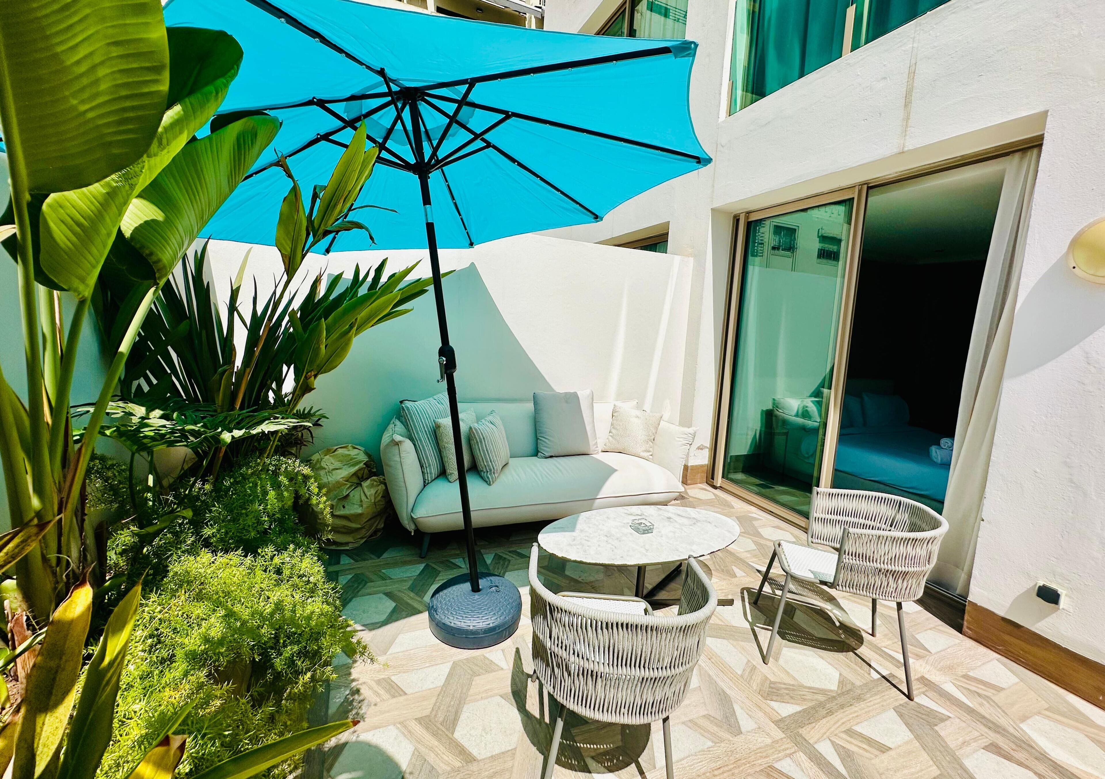 Un espace patio aménagé avec un canapé blanc, une table ronde et des chaises, le tout entouré de plantes en pot et surplombé d’un grand parasol.