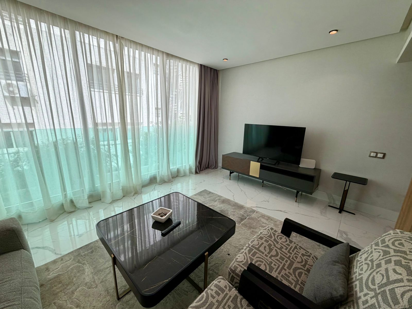 En moderne stue med et glas sofabord, en sofa, et fjernsyn på et stativ og lette gardiner.