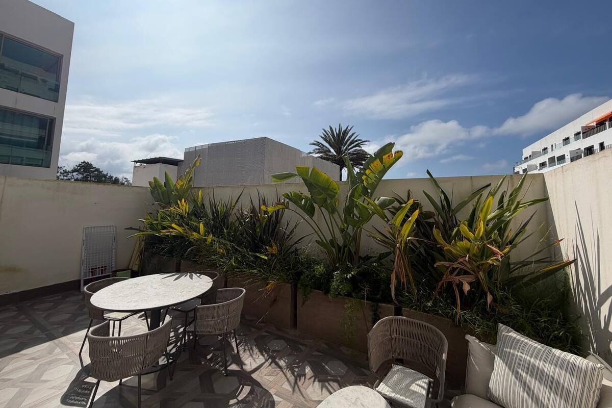 A rooftop patio with a round table and chairs, surrounded by potted plants and a view of buildings.