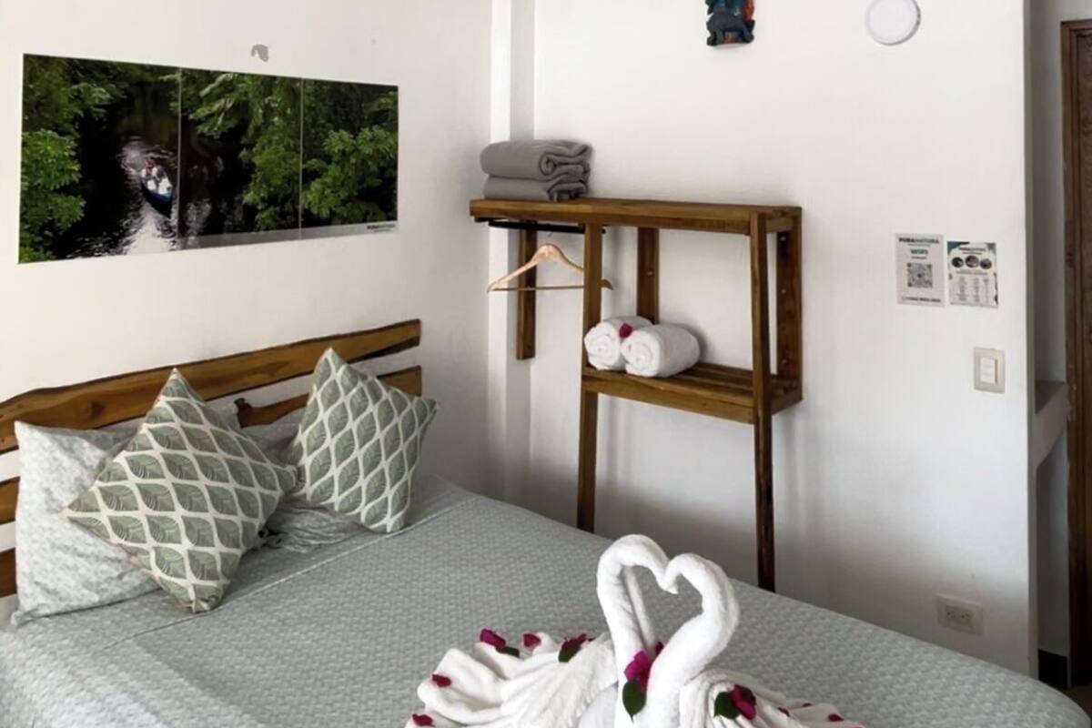 A neatly made bed with a quilted cover, two pillows, a wooden bedside table with towels, and a wall-mounted shelf.