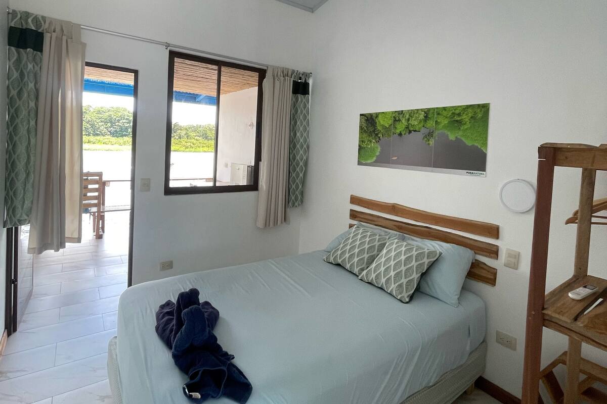 A bedroom with a bed, a wooden headboard, a window with curtains, a painting on the wall, and a view of the outdoors.