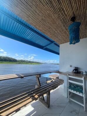 Uma área externa coberta com vista para um corpo d'água, um banco de madeira, uma mesa com cafeteira e um detalhe decorativo de macramé pendurado.