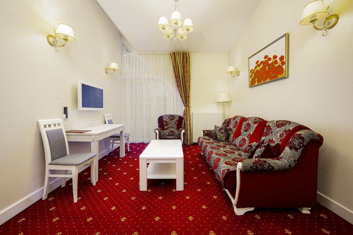A compact living room with a red carpet, white furniture, a television, and a patterned sofa.