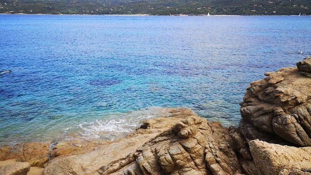 Paisaje costero con aguas azules cristalinas, costa rocosa y montañas a lo lejos.