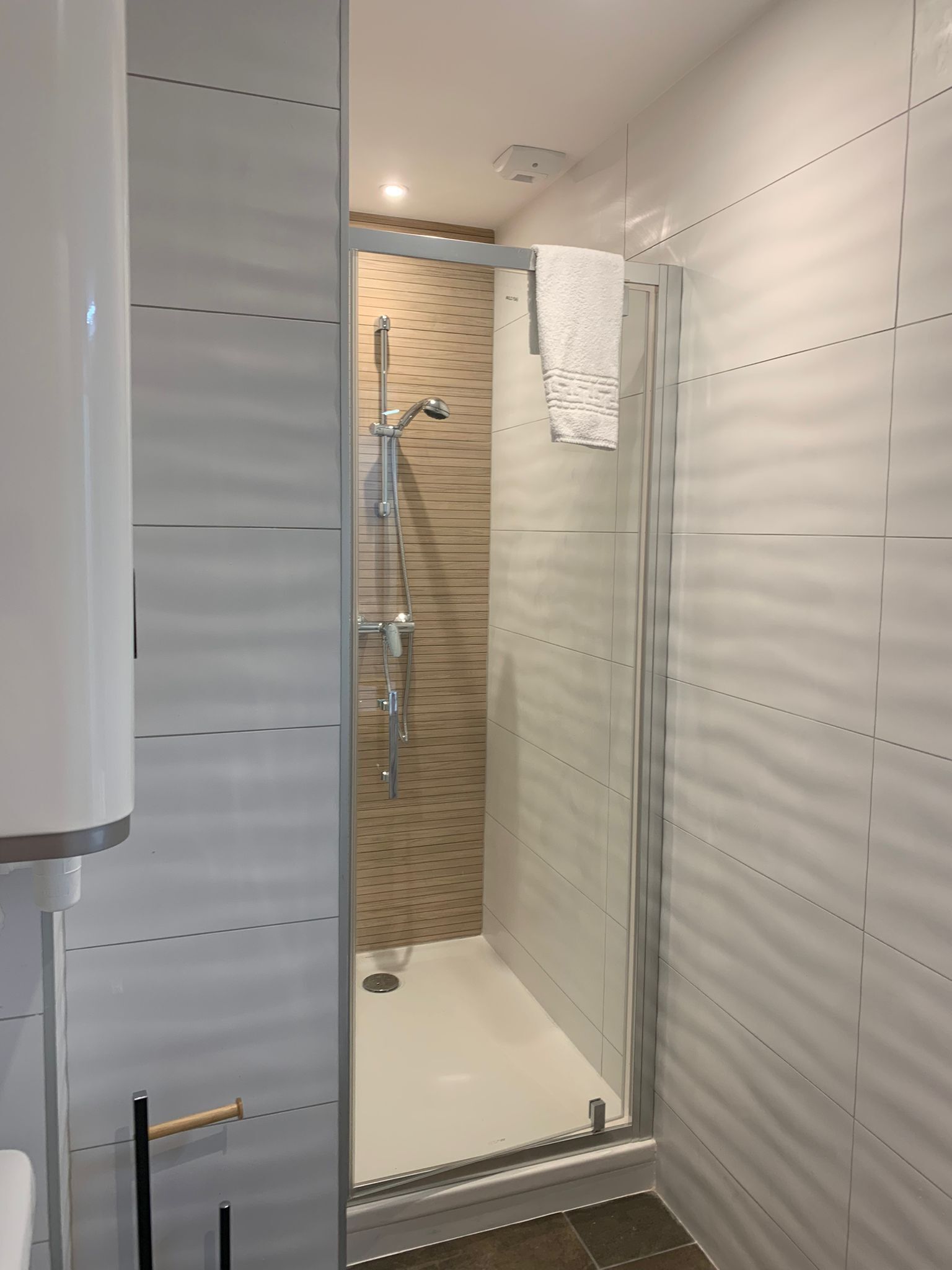 A modern bathroom with a glass shower enclosure, a wooden panel wall, and a white towel hanging on the door.