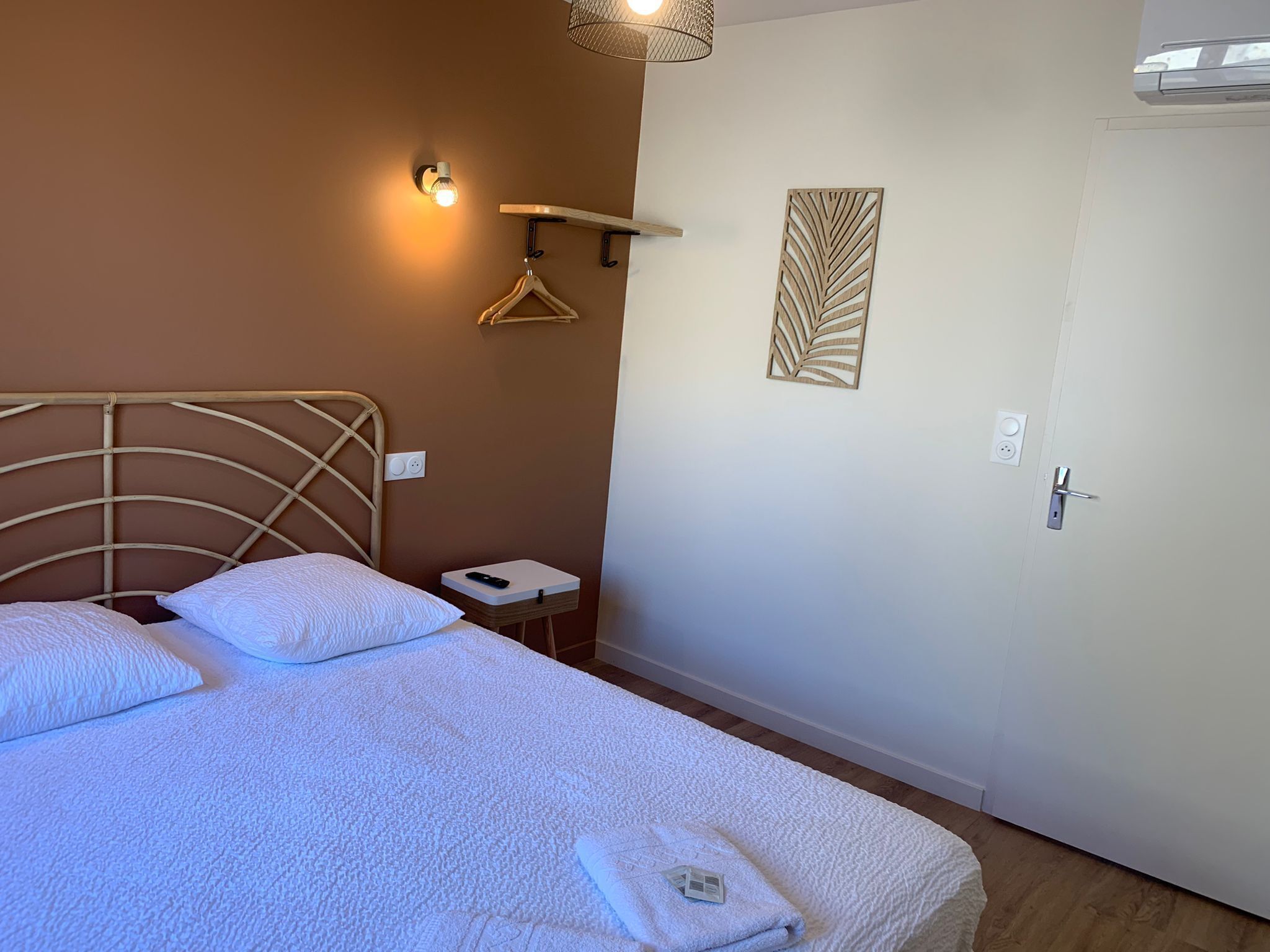 A neatly made bed with a white comforter and pillows, a bedside table with a remote control, a wall-mounted light fixture, and a door with a handle.