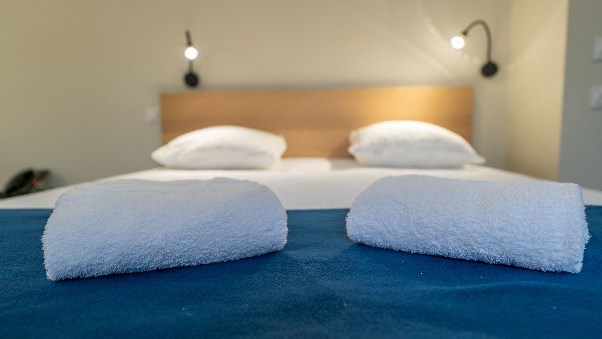 A hotel room with a bed, two white pillows, and two folded white towels on a blue bedspread.