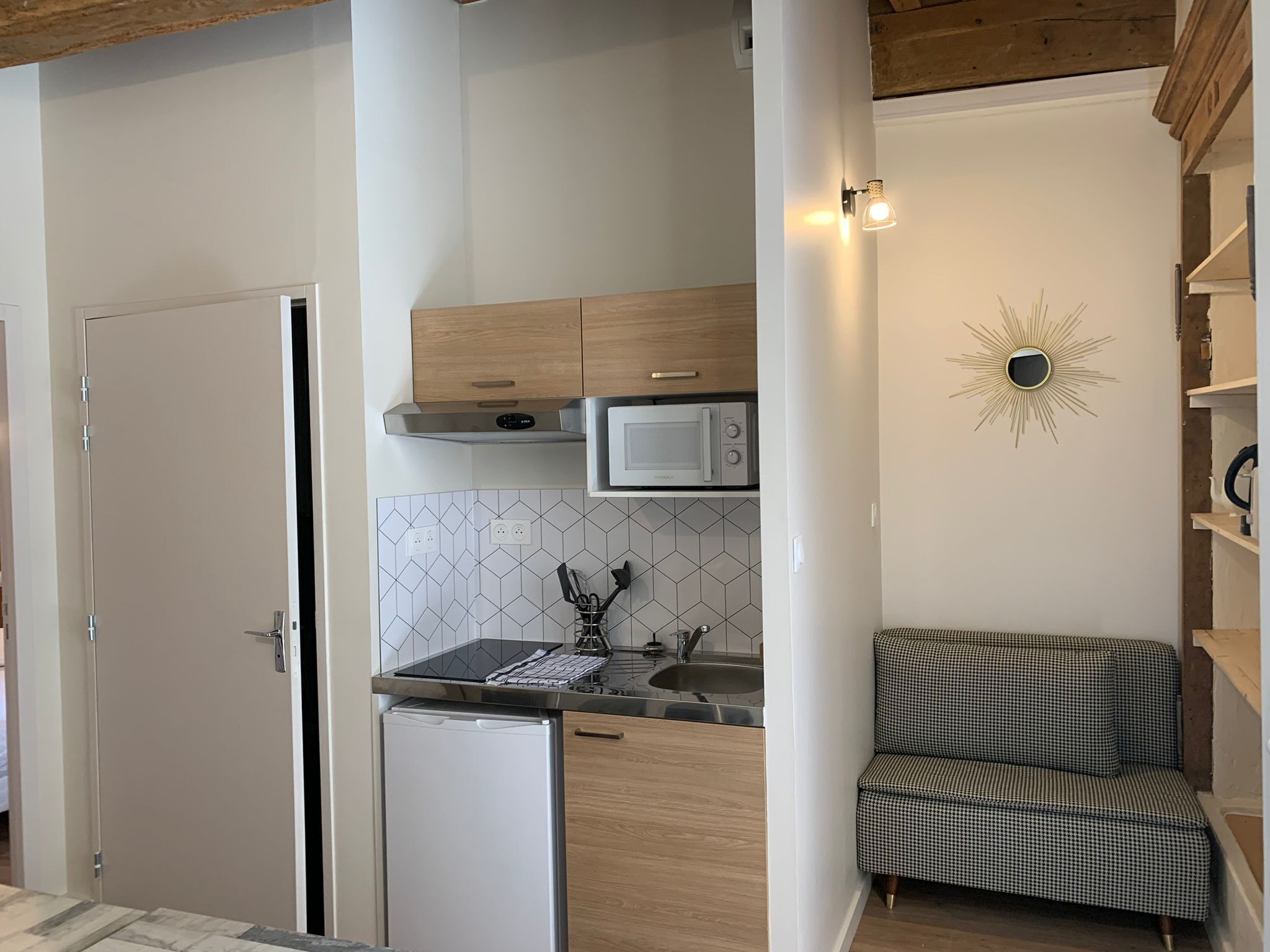 A compact kitchen with a microwave, a small refrigerator, wooden cabinets, and a checkered chair.