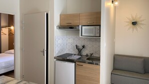 A compact kitchenette with a microwave, a small refrigerator, and a sink. There is a wooden cabinet above the microwave and a houndstooth-patterned sofa to the right. - Hôtel des Docks (Digoin)