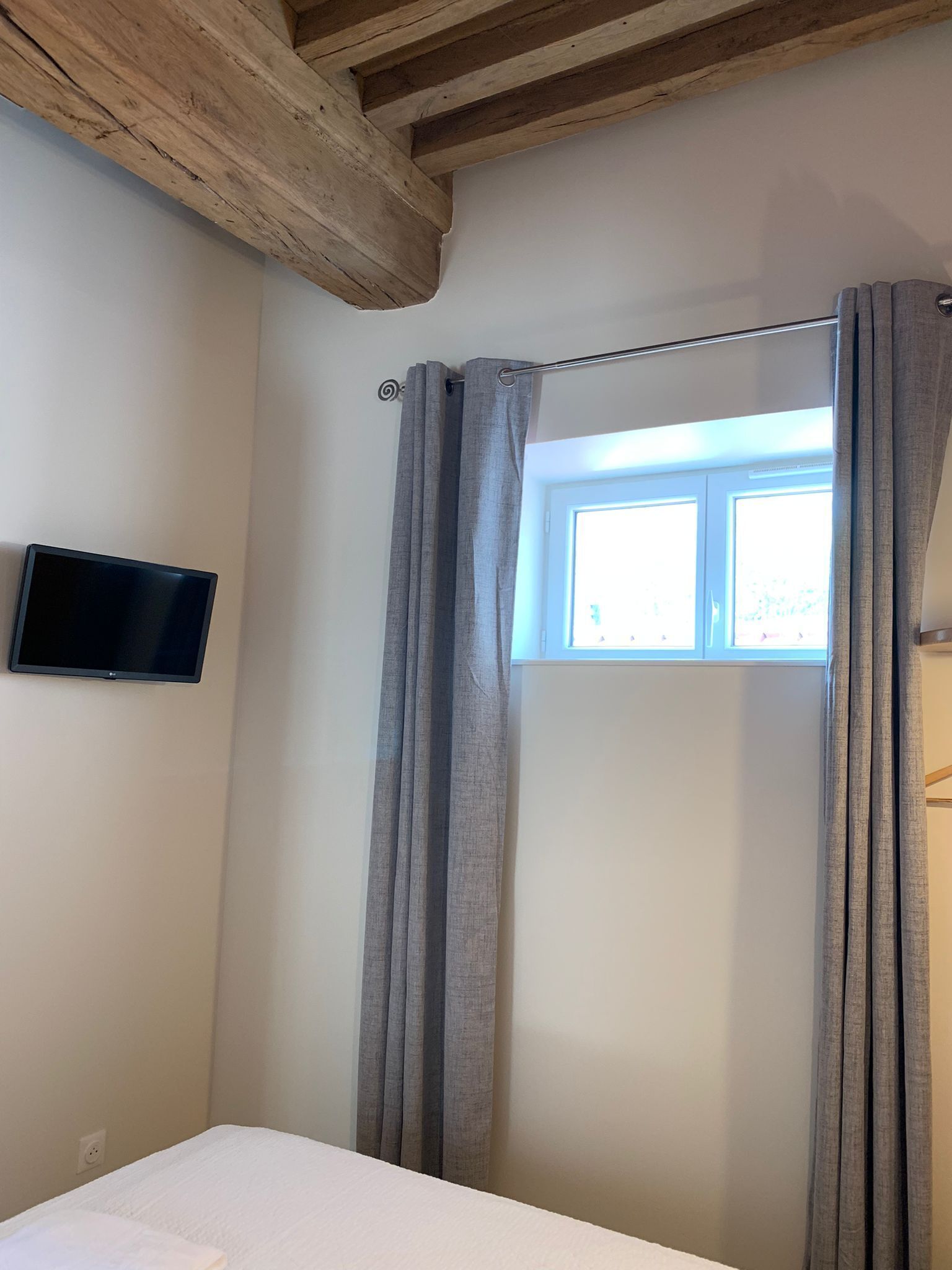 A bedroom with a bed, a window with curtains, a flat-screen TV on the wall, and exposed wooden beams on the ceiling.