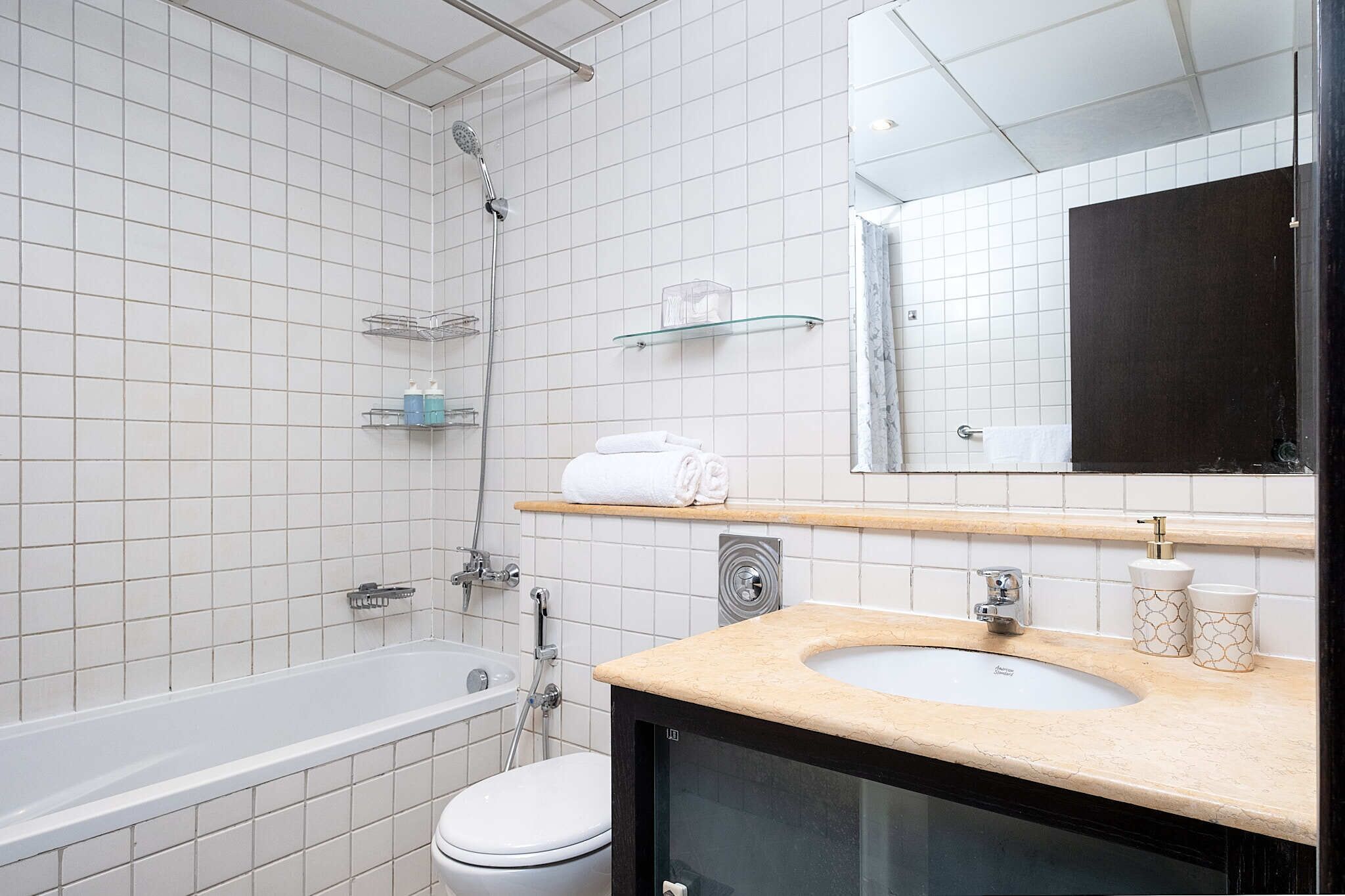 A modern bathroom with a bathtub, toilet, and sink. There are shelves with toiletries and a mirror above the sink.