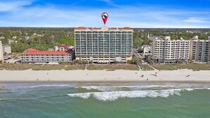 Un hotel frente a la playa con varios pisos, una playa en primer plano y un área boscosa al fondo.