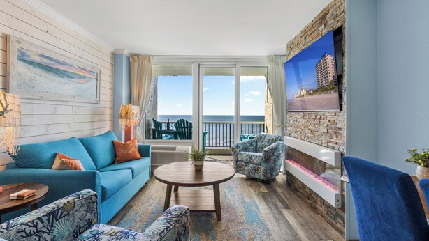 A spacious living room with a blue sofa, patterned armchairs, a round coffee table, and a stone fireplace. A large TV is mounted on the wall, and the room has a view of the ocean. - Carolina Winds 1005-1BR OF Condo in Myrtle Beach (Myrtle Beach)