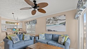 Un salon avec deux canapés bleus, une table basse en bois, un ventilateur de plafond et des œuvres d’art accrochées aux murs.
