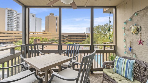 Un balcon avec une table et des chaises, un ventilateur de plafond et une vue sur des immeubles et un parking.