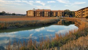 Un hôtel à plusieurs étages, doté d’un toit plat, situé près d’une étendue d’eau bordée de hautes herbes.
